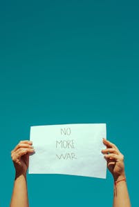 Hands holding a 'No More War' sign against a clear blue sky, symbolizing peace and non-violence.