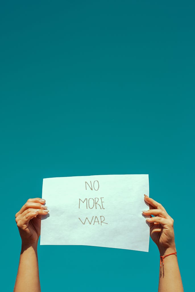 Hands holding a 'No More War' sign against a clear blue sky, symbolizing peace and non-violence.
