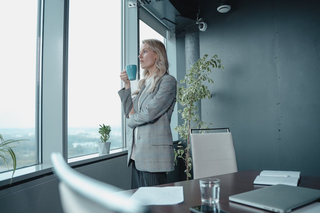 A professional businesswoman enjoys a cup of coffee while overlooking a cityscape from her modern office window.